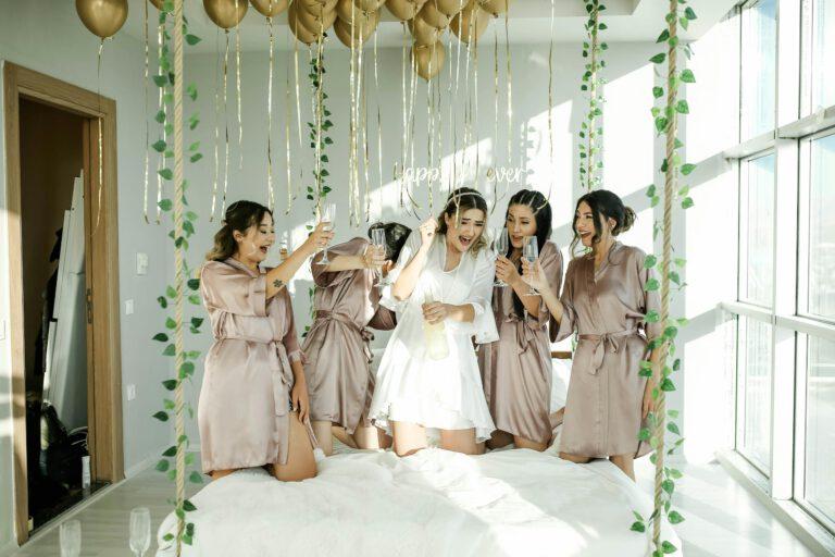 Happy bridal party in satin robes celebrating with champagne and balloons indoors.
