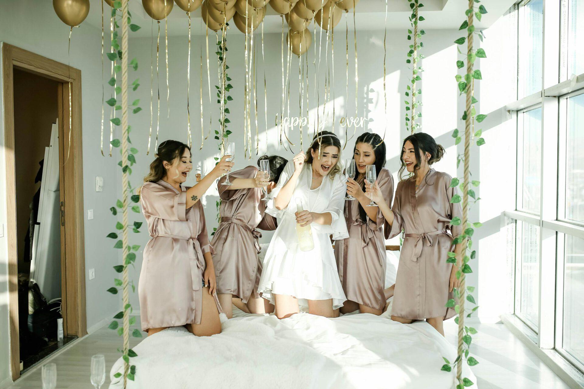 Happy bridal party in satin robes celebrating with champagne and balloons indoors.